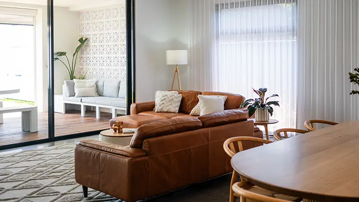 Modern living room with a brown leather sofa, white lamp, indoor plants, and a wooden dining table. A sliding glass door separates the indoors from an outdoor sitting area.