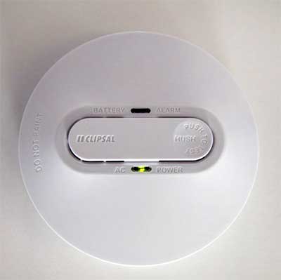 A ceiling-mounted Clipsal smoke alarm with a central control panel showing buttons for hush functions, a battery and alarm status indicator, and an AC power light visible.