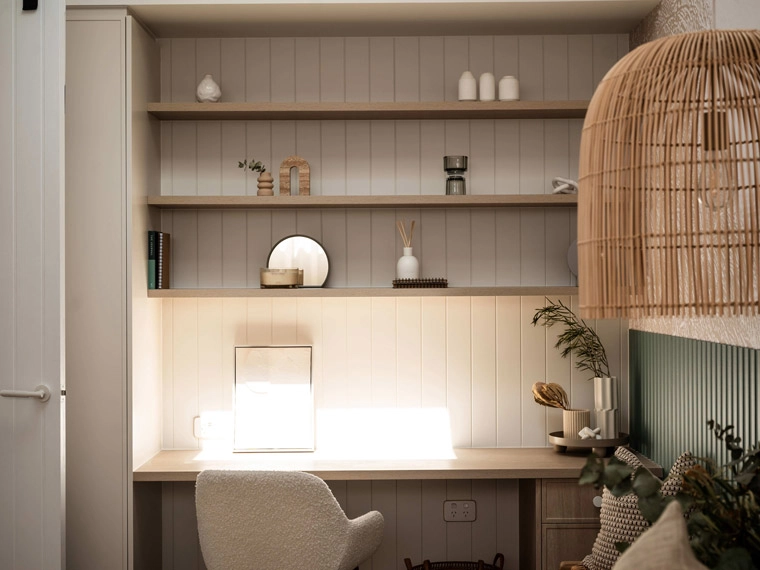 A cozy study corner featuring wall-mounted shelves, a neat work desk with a glowing lamp, and a comfortable chair, decorated in neutral tones.