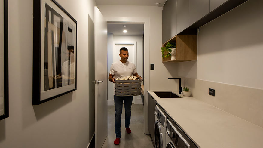 Man carrying his laundry basket in a well-lit laundry room with washing machines and dryer