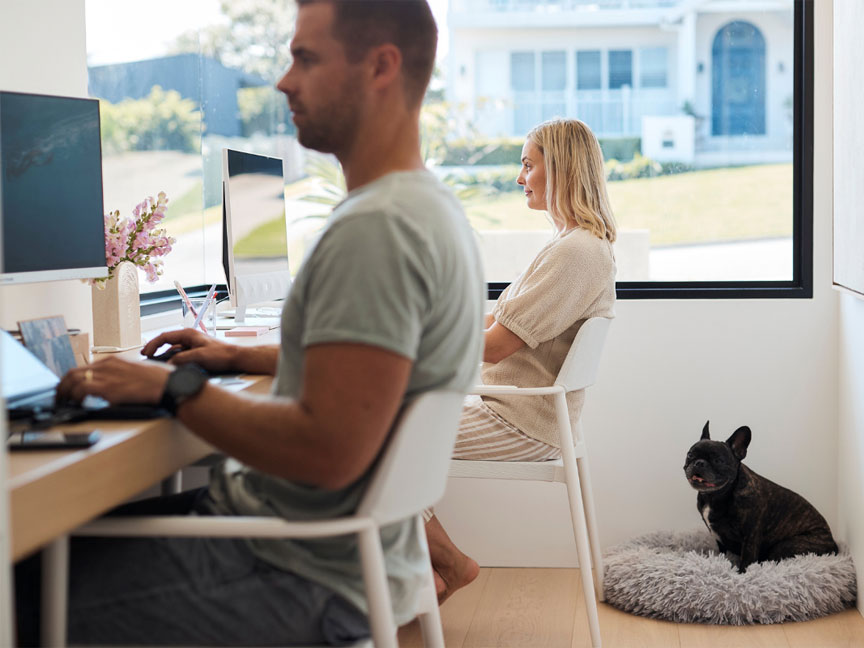 A man and woman seated at a desk, accompanied by a dog resting on a bed nearby