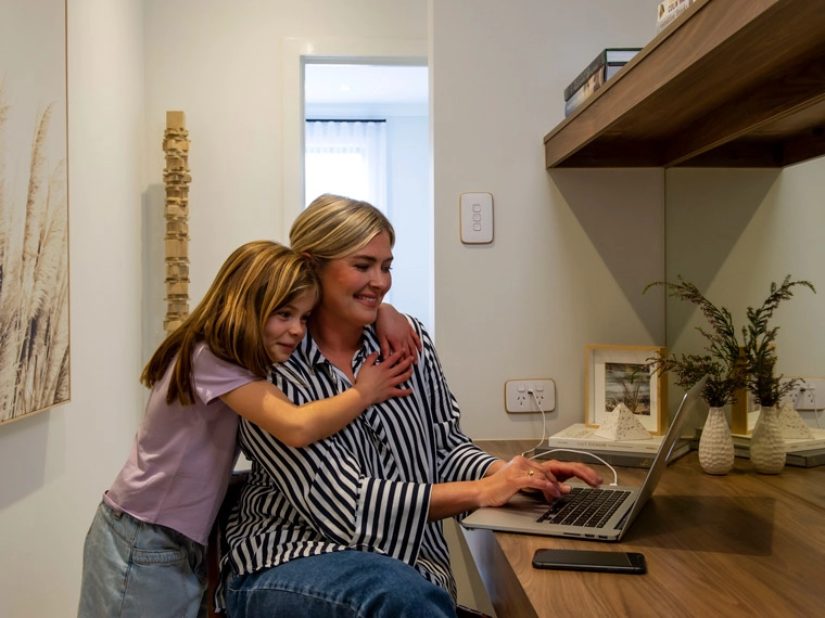 Mother and daughter enjoying time together at a home office, with the mother using a laptop and the daughter hugging her from behind, in a cozy room with stylish interior decor.